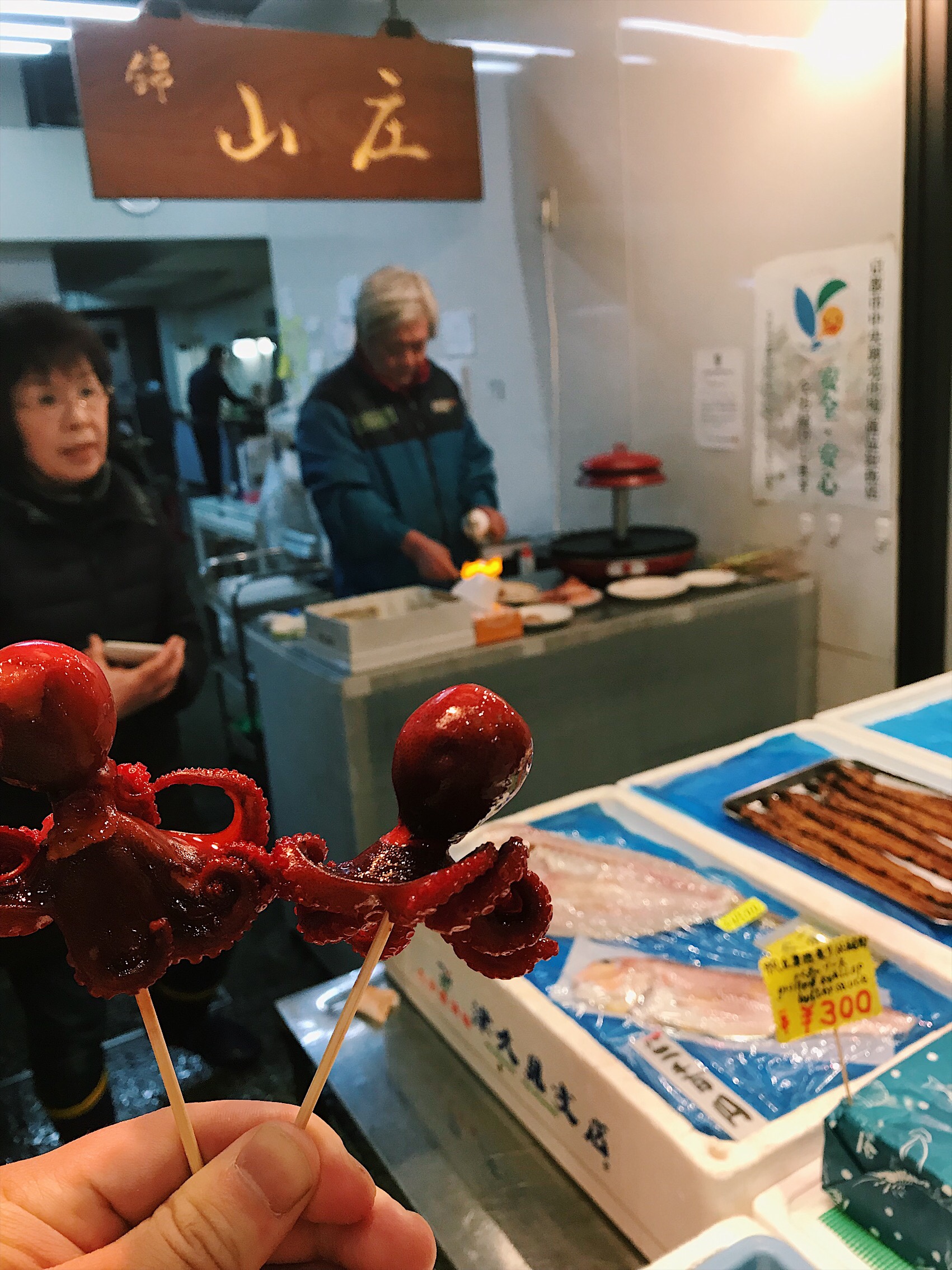 two small red octopi on skewers with Japanese man and woman in background