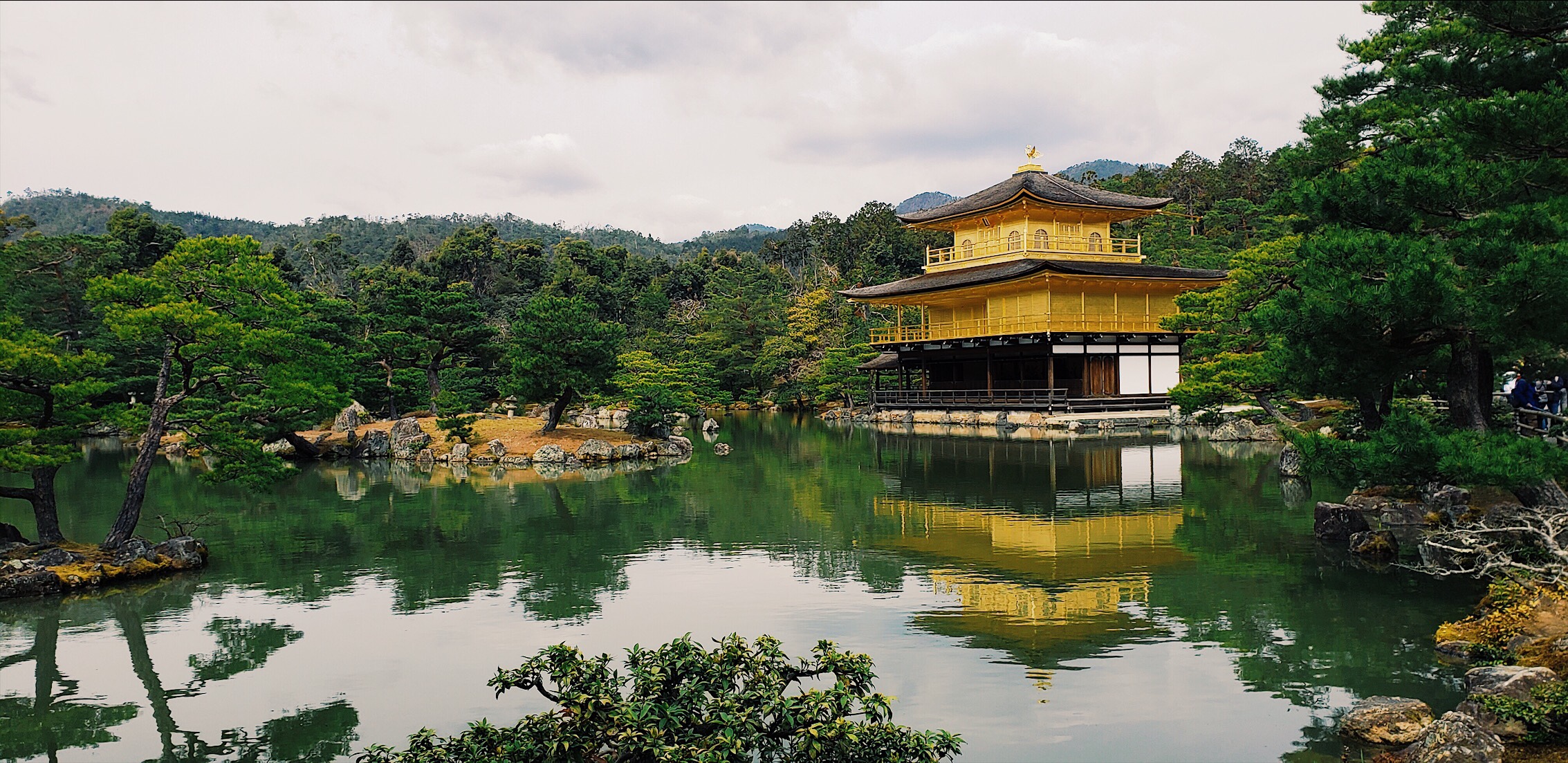 Three story golden temple sitting in front of pond, surrounded by rocky Zen gardens and trees